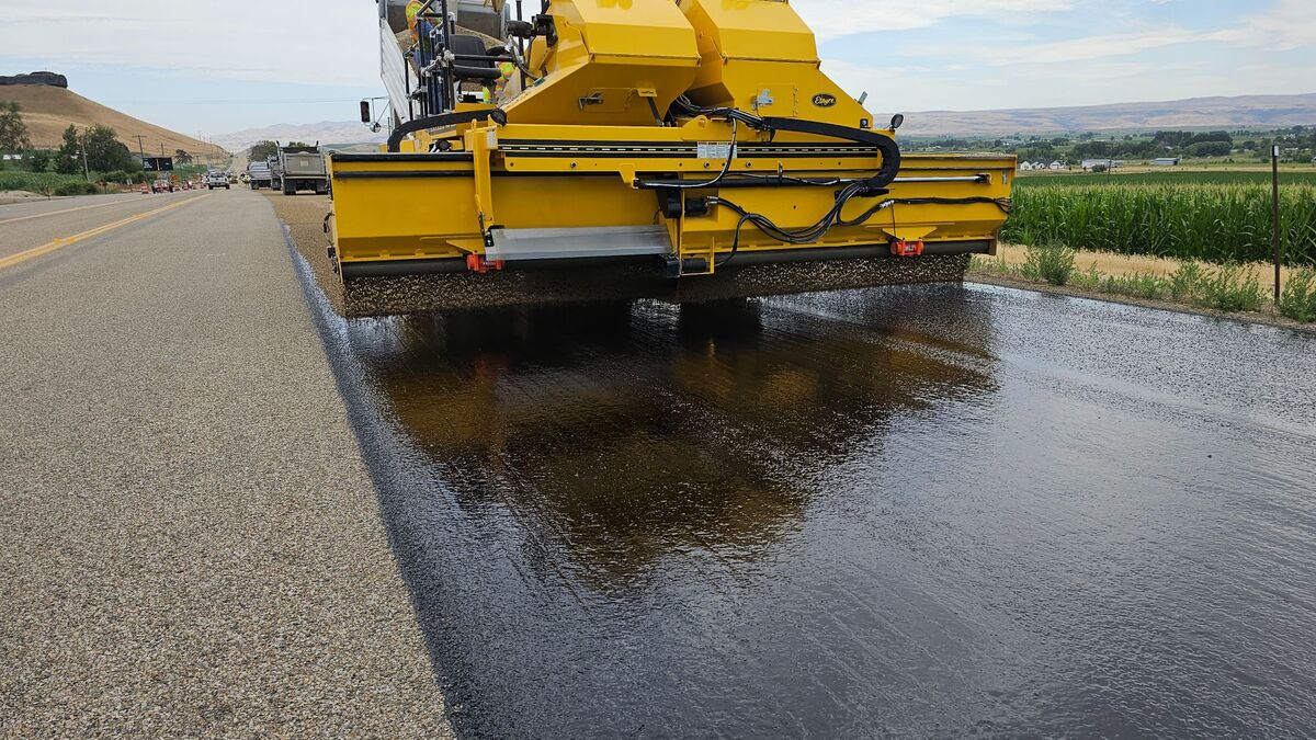 An asphalt paver laying a fresh black mat on a long, flat road past open farmland